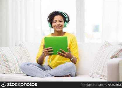 people, technology and leisure concept - happy african american young woman sitting on sofa with tablet pc computer and headphones listening to music at home