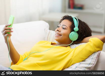 people, technology and leisure concept - happy african american young woman sitting on sofa with smartphone and headphones listening to music at home