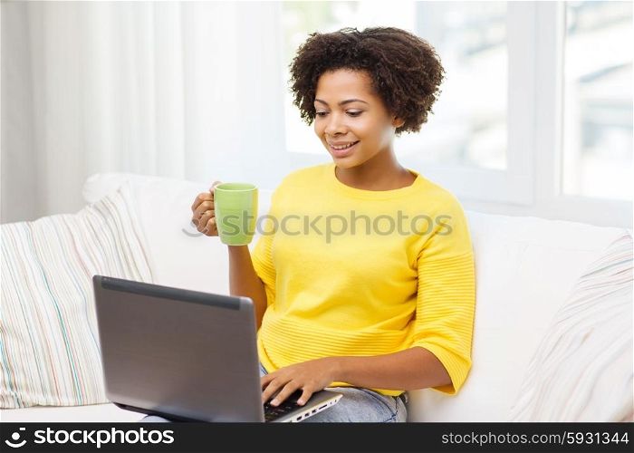 people, technology and leisure concept - happy african american young woman sitting on sofa with laptop computer and drinking tea from cup at home