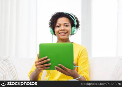 people, technology and leisure concept - happy african american young woman sitting on sofa with tablet pc computer and headphones listening to music at home