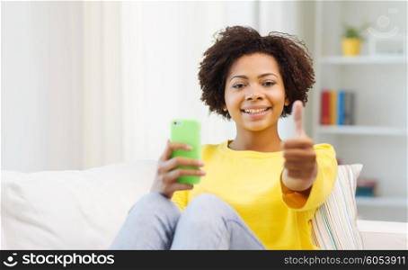 people, technology and leisure concept - happy african american young woman sitting on sofa with smartphone and showing thumbs up at home