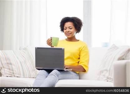 people, technology and leisure concept - happy african american young woman sitting on sofa with laptop computer and drinking tea from cup at home
