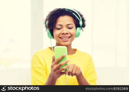 people, technology and leisure concept - happy african american young woman sitting on sofa with smartphone and headphones listening to music at home