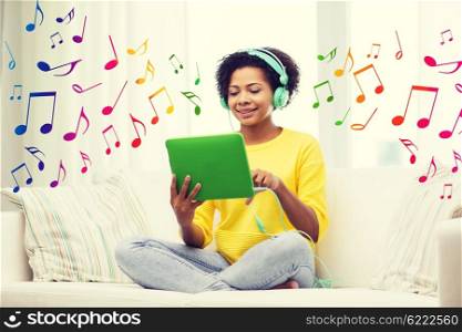 people, technology and leisure concept - happy african american young woman sitting on sofa with tablet pc computer and headphones listening to music at home over notes background