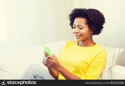 people, technology and leisure concept - happy african american young woman sitting on sofa with smartphone at home