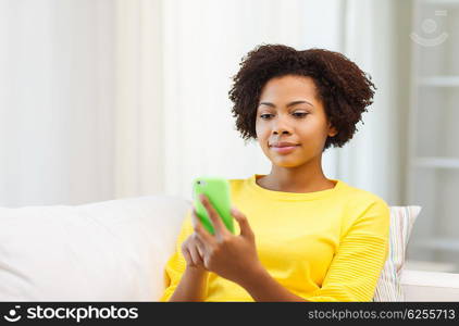 people, technology and leisure concept - happy african american young woman sitting on sofa with smartphone at home