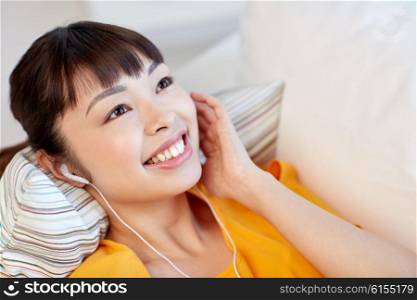 people, technology and leisure concept - happy asian young woman lying on sofa with earphones listening to music at home