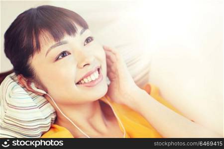 people, technology and leisure concept - happy asian young woman lying on sofa with earphones listening to music at home. happy asian woman with earphones listening music