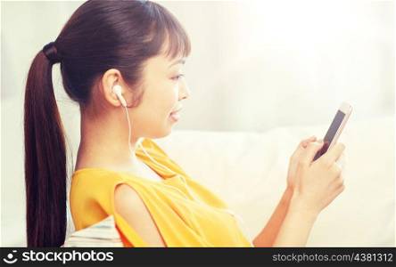 people, technology and leisure concept - happy asian young woman sitting on sofa with smartphone and earphones listening to music at home. happy asian woman with smartphone and earphones. happy asian woman with smartphone and earphones