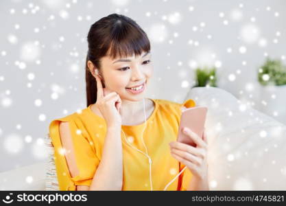 people, technology and leisure concept - happy asian young woman sitting on sofa with smartphone and earphones listening to music at home over snow