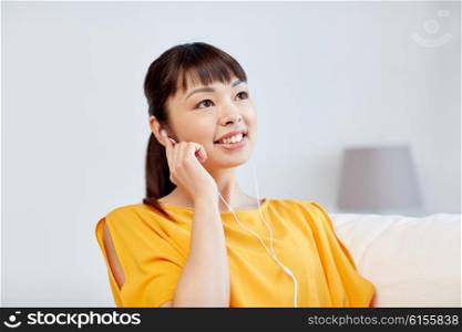 people, technology and leisure concept - happy asian young woman sitting on sofa with earphones listening to music at home