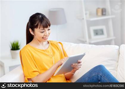 people, technology and leisure concept - happy asian young woman sitting on sofa with tablet pc computer and earphones listening to music at home