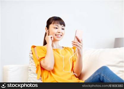 people, technology and leisure concept - happy asian young woman sitting on sofa with smartphone and earphones listening to music at home