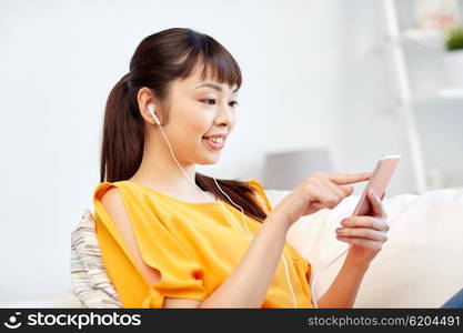 people, technology and leisure concept - happy asian young woman sitting on sofa with smartphone and earphones listening to music at home