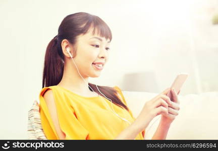 people, technology and leisure concept - happy asian young woman sitting on sofa with smartphone and earphones listening to music at home. happy asian woman with smartphone and earphones. happy asian woman with smartphone and earphones