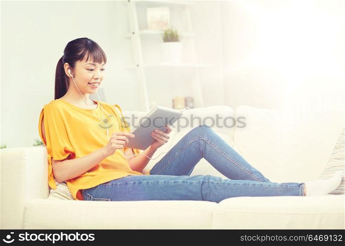 people, technology and leisure concept - happy asian young woman sitting on sofa with tablet pc computer and earphones listening to music at home. happy asian woman with tablet pc and earphones