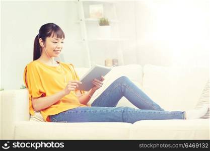 people, technology and leisure concept - happy asian young woman sitting on sofa with tablet pc computer and earphones listening to music at home. happy asian woman with tablet pc and earphones
