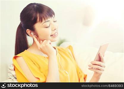 people, technology and leisure concept - happy asian young woman sitting on sofa with smartphone and earphones listening to music at home. happy asian woman with smartphone and earphones