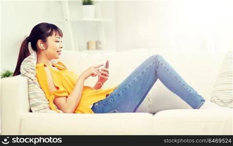 people, technology and leisure concept - happy asian young woman sitting on sofa with smartphone and earphones listening to music at home. happy asian woman with smartphone and earphones