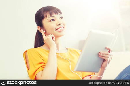 people, technology and leisure concept - happy asian young woman sitting on sofa with tablet pc computer and earphones listening to music at home. happy asian woman with tablet pc and earphones