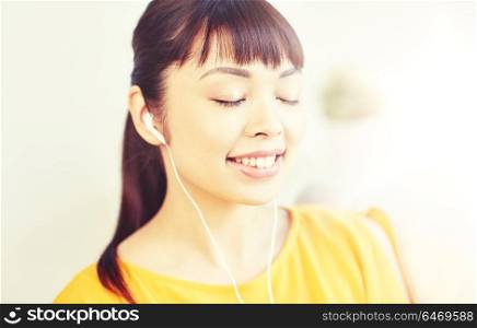 people, technology and leisure concept - happy asian young woman sitting on sofa with earphones listening to music at home. happy asian woman with earphones listening music