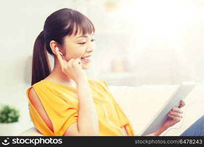 people, technology and leisure concept - happy asian young woman sitting on sofa with tablet pc computer and earphones listening to music at home. happy asian woman with tablet pc and earphones. happy asian woman with tablet pc and earphones