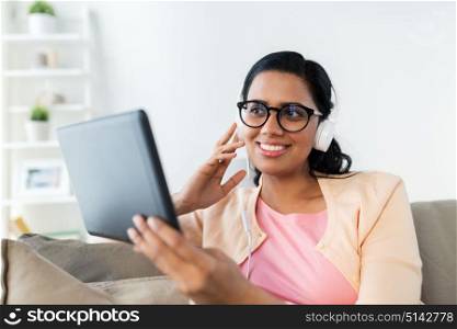 people, technology and leisure concept - happy indian young woman sitting on sofa with tablet pc computer and headphones listening to music at home. happy woman with tablet pc and headphones at home