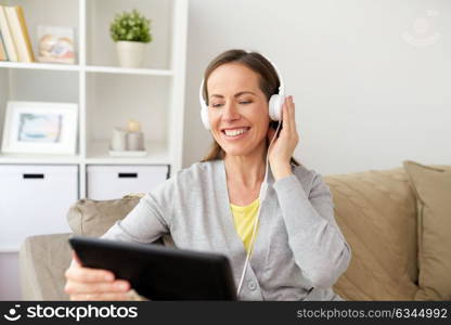 people, technology and leisure concept - happy middle-aged woman sitting on sofa with tablet pc computer and headphones listening to music at home. happy woman with tablet pc and headphones at home