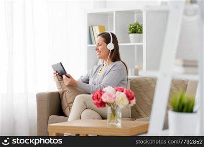 people, technology and leisure concept - happy middle-aged woman sitting on sofa with tablet pc computer and headphones listening to music at home. happy woman with tablet pc and headphones at home