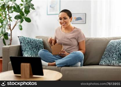 people, technology and leisure concept - happy smiling african american young woman eating takeaway food with chopsticks and watching movie on tablet pc computer at home. african woman with tablet pc eating takeaway food