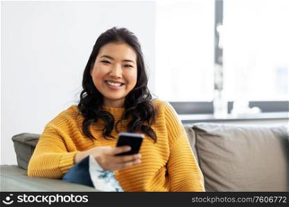 people, technology and leisure concept - happy smiling asian young woman in yellow sweater with smartphone sitting on sofa at home. happy asian young woman with smartphone at home