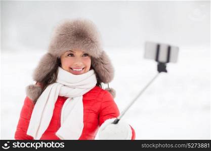 people, technology and leisure concept - happy smiling woman in winter fur hat taking picture by smartphone on selfie stick outdoors. happy woman with selfie stick outdoors in winter