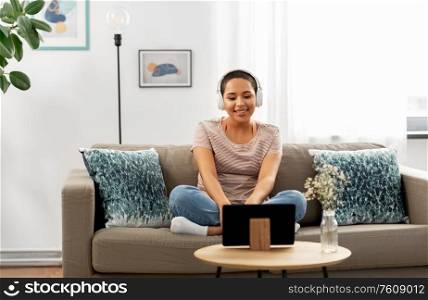 people, technology and leisure concept - happy smiling young african american woman in headphones sitting on sofa and listening to music or watching movie on tablet pc computer at home. woman with headphones and tablet computer at home