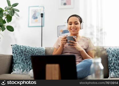 people, technology and leisure concept - happy smiling young african american woman in headphones sitting on sofa with tablet pc computer, drinking coffee and watchin movie at home. woman with tablet pc drinking coffee at home