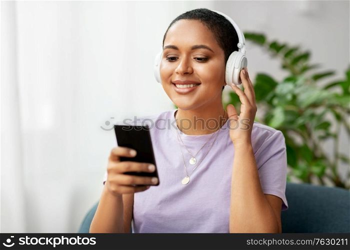 people, technology and leisure concept - happy smiling young african american woman in glasses with smartphone and headphones sitting in chair and listening to music at home. woman with smartphone listening to music at home