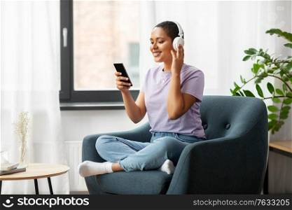 people, technology and leisure concept - happy smiling young african american woman in glasses with smartphone and headphones sitting in chair and listening to music at home. woman with smartphone listening to music at home