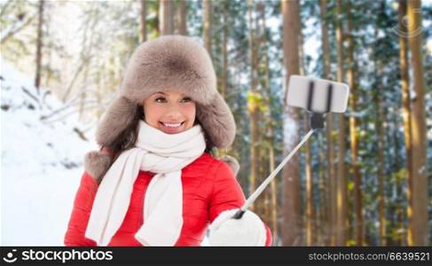 people, technology and leisure concept - happy woman in fur hat taking picture by smartphone selfie stick over winter forest background. happy woman taking selfie over winter forest