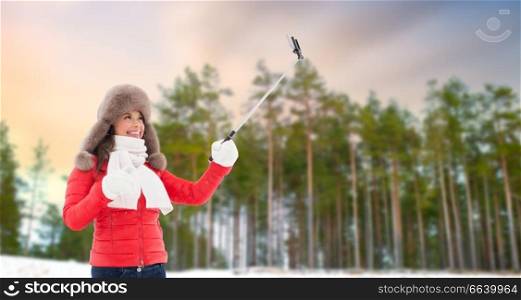 people, technology and leisure concept - happy woman in fur hat taking picture by smartphone selfie stick over winter forest background. happy woman taking selfie over winter forest