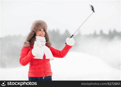 people, technology and leisure concept - happy woman in winter fur hat taking picture by smartphone on selfie stick outdoors. happy woman with selfie stick outdoors in winter