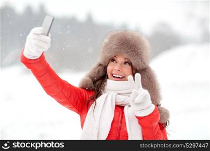 people, technology and leisure concept - happy woman in winter fur hat taking selfie by smartphone outdoors. happy woman taking selfie outdoors in winter