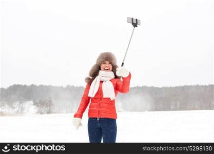 people, technology and leisure concept - happy woman in winter fur hat taking picture by smartphone on selfie stick outdoors. happy woman with selfie stick outdoors in winter