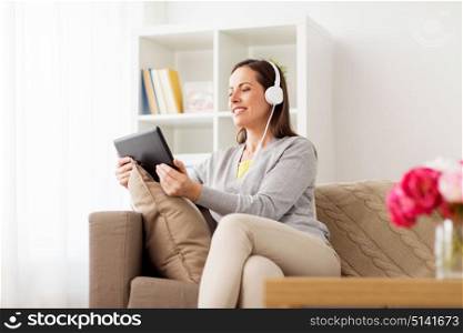 people, technology and leisure concept - happy woman sitting on sofa with tablet pc computer and headphones listening to music at home. happy woman with tablet pc and headphones at home