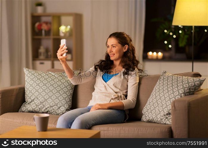 people, technology and leisure concept - happy woman taking selfie with smartphone at home in evening. happy woman taking selfie with smartphone at home