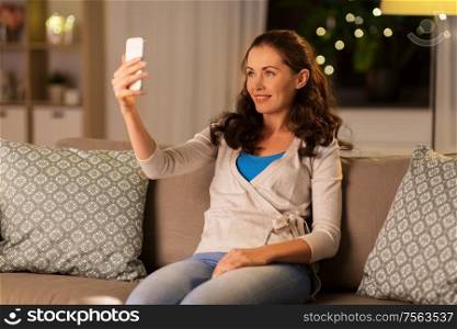 people, technology and leisure concept - happy woman taking selfie with smartphone at home in evening. happy woman taking selfie with smartphone at home