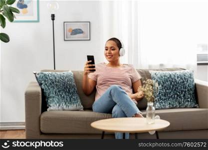 people, technology and leisure concept - happy young african american woman in headphones with smartphone sitting on sofa and listening to music at home. woman with smartphone listening to music at home