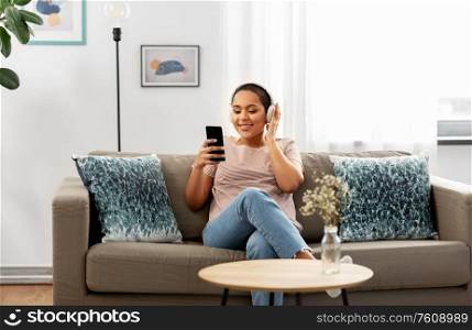 people, technology and leisure concept - happy young african american woman in glasses with smartphone and headphones sitting on sofa and listening to music at home. woman with smartphone listening to music at home