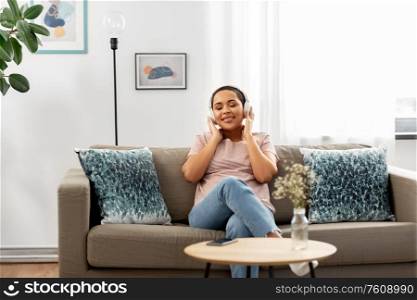 people, technology and leisure concept - happy young african american woman in glasses with headphones and smartphone sitting on sofa and listening to music at home. woman with headphones listening to music at home