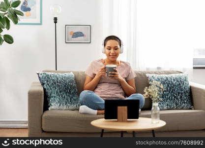 people, technology and leisure concept - happy young african american woman in headphones sitting on sofa, drinking coffee and watching movie on tablet pc computer at home. woman with headphones and tablet pc drinking tea