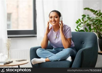people, technology and leisure concept - happy young african american woman in headphones sitting in chair and listening to music at home. woman in headphones listening to music at home
