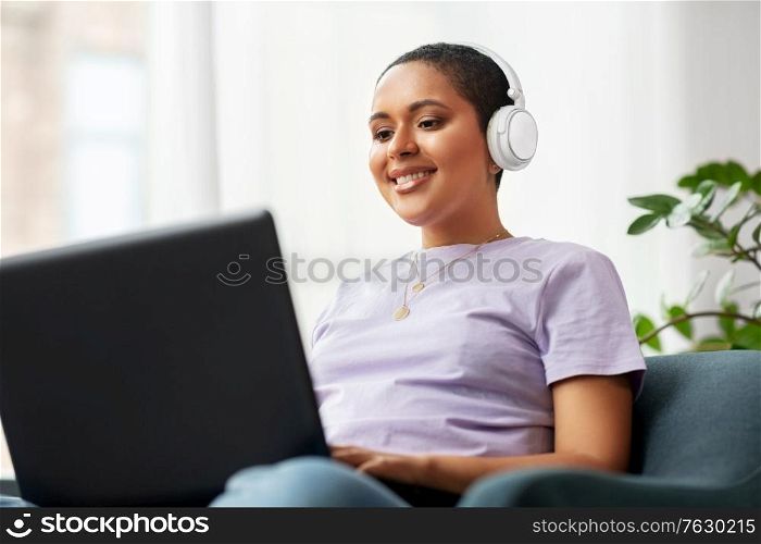 people, technology and leisure concept - happy young african american woman in headphones with laptop computer listening to music at home. woman with laptop listening to music at home